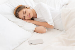 Young female covered by white blanket on bed, enjoying muisc via wireless headphones Young female covered by white blanket on bed, enjoying muisc via wireless headphones